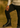Man wearing a beige sweater and black pants standing on a dirt path with greenery in the background