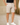Man wearing beige cotton chino shorts and white sneakers, standing in an elegant room with marble floors and beige paneled walls