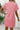 Person wearing a pink textured dress standing against a white wall with a plant in the background