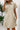 Woman wearing a beige dress with a textured pattern, holding a phone, against a white wall