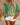 Woman wearing a green and yellow patterned top with white shorts, holding a straw bag