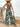 Woman wearing a green leaf-patterned jumpsuit with a straw hat and sandals, standing in a casual setting