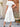 Woman wearing a white dress standing outdoors near a wooden door and wicker chair