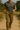 Australian male model in motion wearing khaki cargo pants with beige belt, walking on a sunlit dirt path surrounded by greenery. Captured in natural light with full view of the pants and rugged outdoor setting
