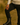 Man wearing a beige sweater and black pants standing on a dirt path with greenery in the background