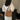 Back view of a woman in a white embroidered dress wearing a brown woven hobo shoulder bag with netted center panel and leather strap, standing indoors near a wooden chair