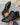 Black ankle boots with brown soles held by a hand on a gray surface