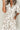 Close-up of a woman wearing a white midi dress with tan floral print, black buttons, puff sleeves, and side pockets, accessorized with gold bracelets and a neutral manicure