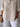 Person wearing a beige button-up shirt and white pants against a light wooden panel background