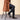 Person wearing a brown coat, dark pants, and black shoes sitting on a stool against a beige background