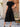 Woman wearing a black dress standing outdoors near a wooden door and wicker chair
