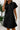 Woman wearing a black dress with a collar, standing against a white wall with a plant in the background