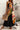 Woman wearing a black dress with colorful abstract patterns, holding a woven bag against a concrete wall