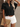 Woman wearing a black ruffled blouse, white shorts, and sunglasses, holding a straw clutch