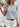 Woman wearing a floral top and white shorts with a straw hat, standing against a white wall