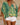Woman wearing a green and yellow patterned top with white shorts, holding a straw bag