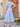 Woman wearing a light blue dress standing outdoors near a wooden door, back view