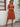 Woman wearing a rust-colored dress with a straw hat and sunglasses, standing against a white wall