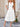 Woman wearing a white dress standing outdoors with a wicker chair and decorative wall in the background