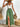Woman wearing a white top and green skirt with a straw hat and bag, standing in a rustic setting
